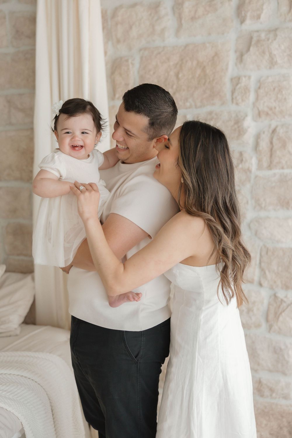 Happy family with father holding smiling baby girl while mother kisses his cheek in bright white room.