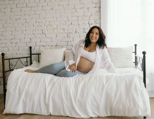 Expectant mother in white shirt and jeans lounges on white bedding against brick wall background.