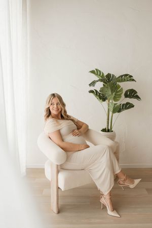 Pregnant woman in beige outfit sitting in white chair beside monstera plant in bright minimalist studio.