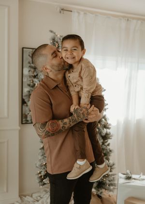 A parent with tattoos hugs and kisses their child while wearing a brown sweater in a bright indoor setting.