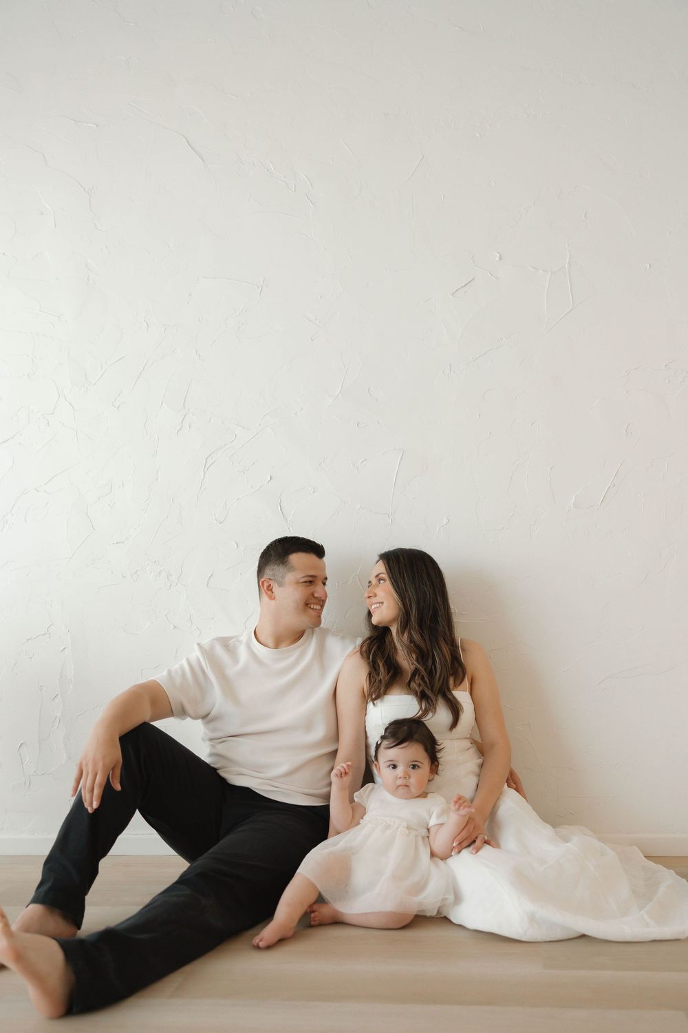 A happy young family with a baby girl sitting on a hardwood floor against a white textured wall.
