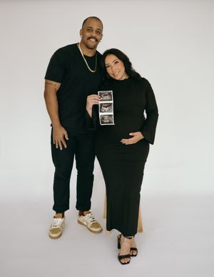 Couple in black outfits poses with ultrasound photos against white background.