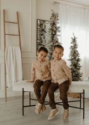 Children in matching beige outfits sit playfully on black bench in bright studio setting.