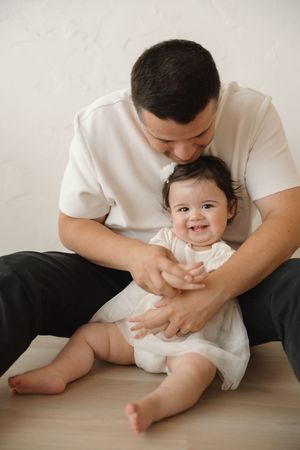 A father tenderly kisses his smiling baby girl while holding her on a light wood floor.