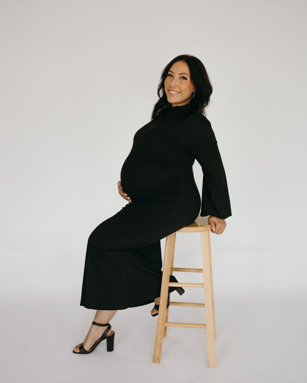 Woman in black dress poses on wooden stool for maternity photo.
