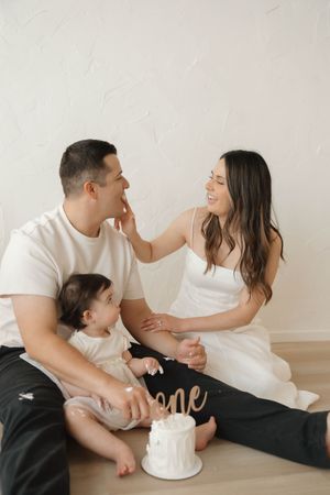 Happy family celebrates baby's first birthday with a white smash cake on a wood floor.