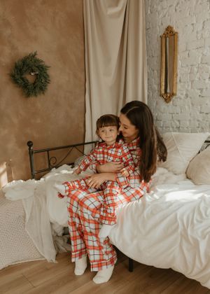 Family wearing matching plaid pajamas relaxes on white bedding against brick wall.