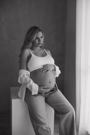 Black and white maternity portrait of pregnant woman in casual attire by window at a studio in Huntington Beach