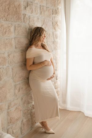 Pregnant woman in beige off-shoulder dress cradles baby bump, leaning against stone wall near sheer curtained window.