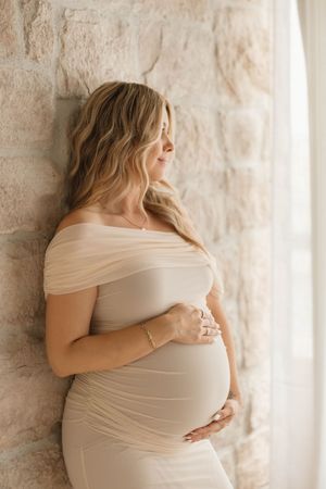 Pregnant woman in beige off-shoulder dress cradling baby bump, leaning against stone wall near window.