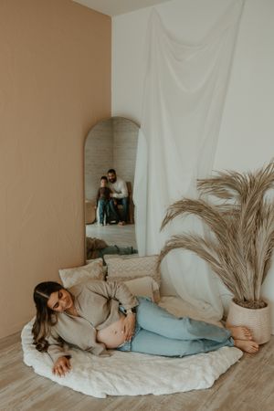 A maternity photo featuring a pregnant woman relaxing on a white blanket with pampas grass decor.