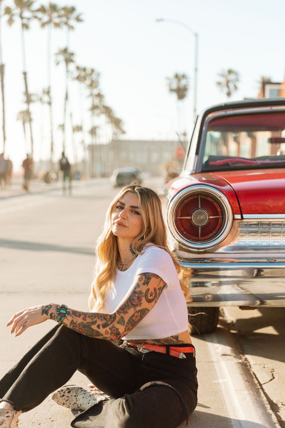 Tattooed woman in casual attire sitting beside vintage red car on palm tree lined street during sunset.