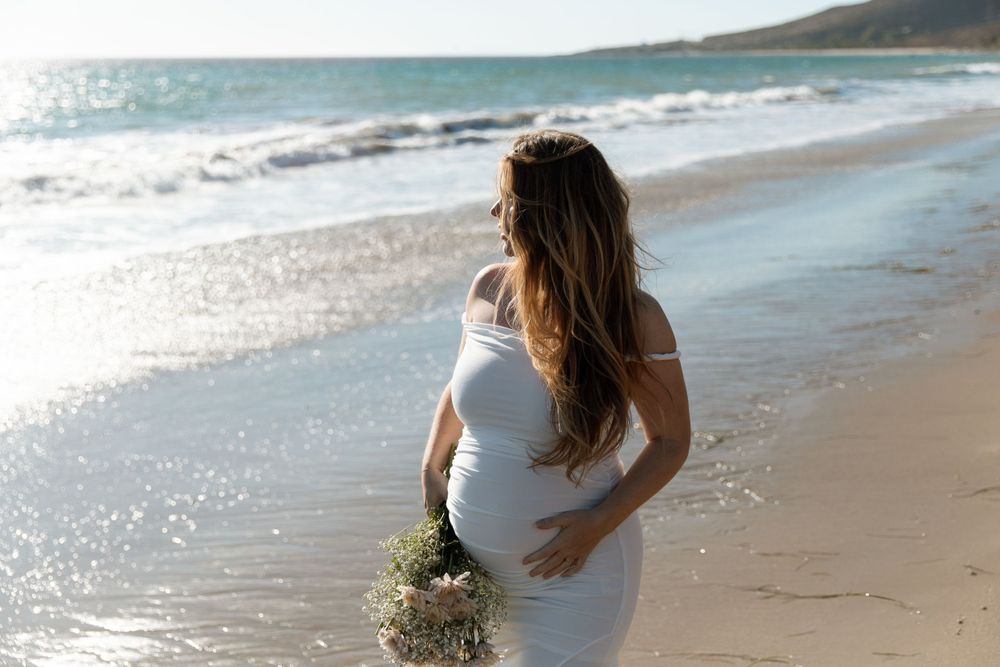 Expectant mother in white dress holds bouquet while standing on sunny beach at golden hour.