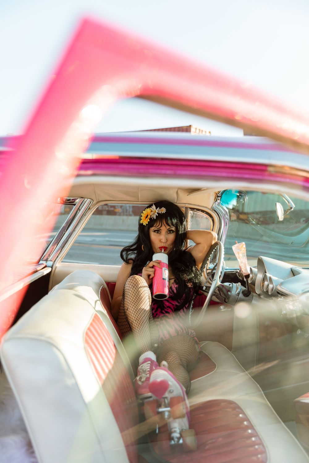 View through car window of retro car interior with person sipping pink drink while wearing flower crown and vintage outfit.