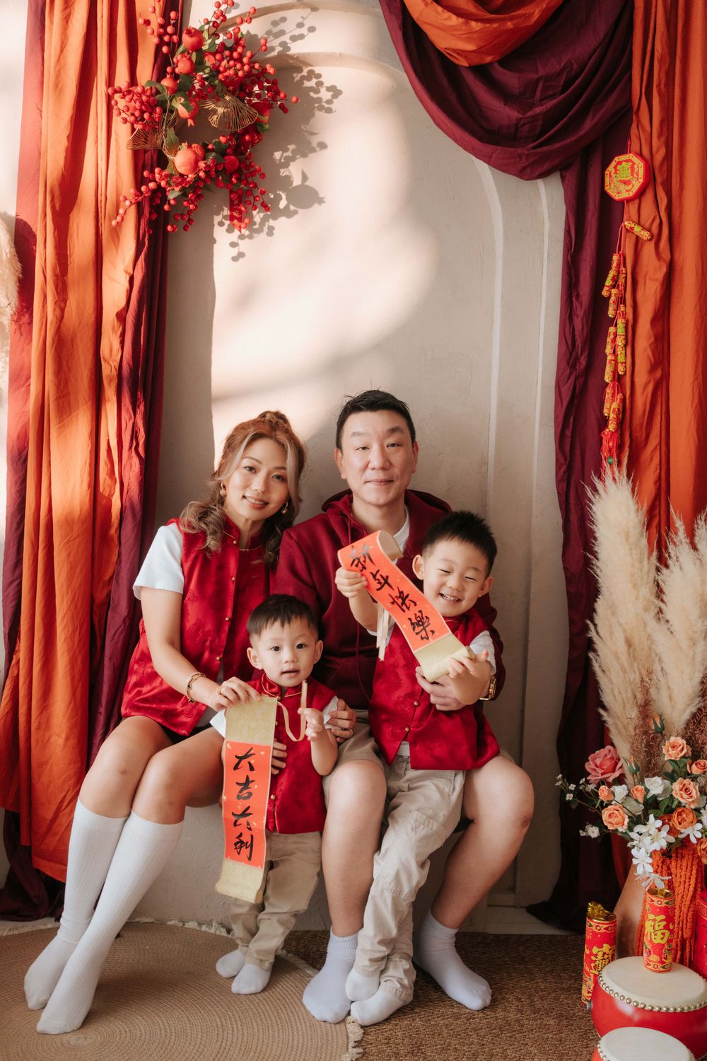 Family wearing red holiday outfits poses together with traditional Chinese New Year decorations and banners.