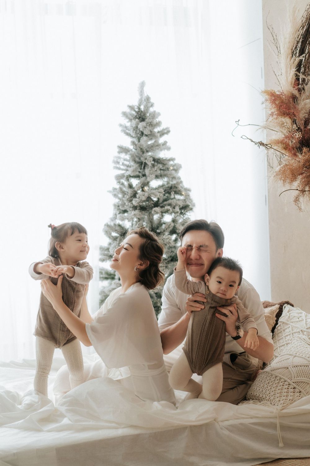 Joyful family playtime on bed with two children in front of festive Christmas tree.