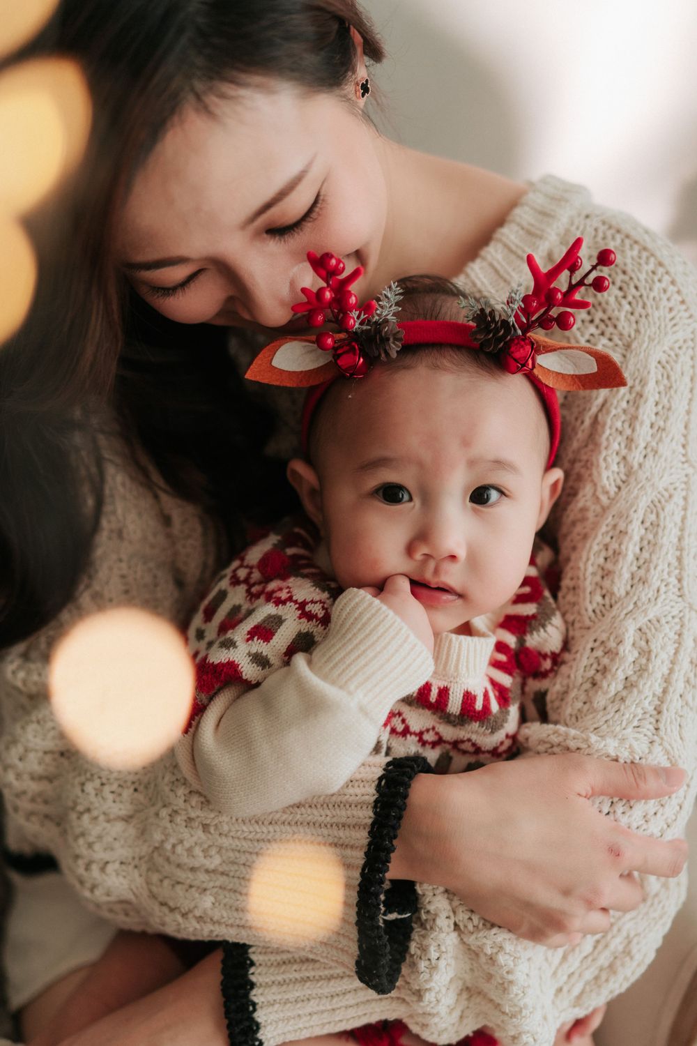 Mother and baby in matching knit sweaters share tender moment with festive reindeer accessories.