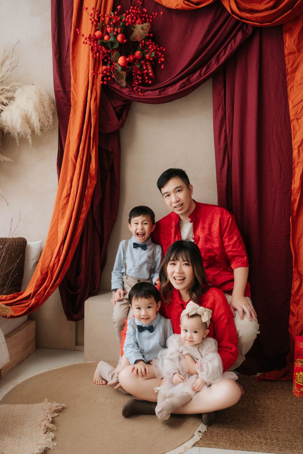 Family portrait with red drapery backdrop featuring children in gray outfits and baby in white.