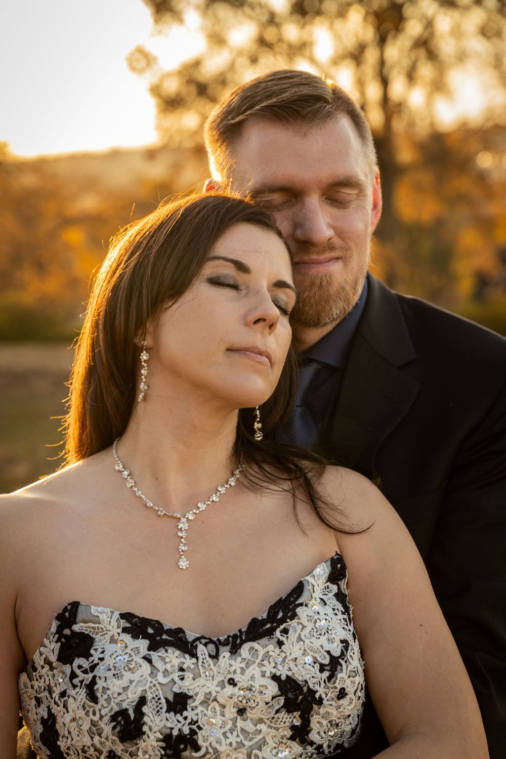 A romantic couple embraces in golden sunset light while wearing formal attire with delicate lace details.