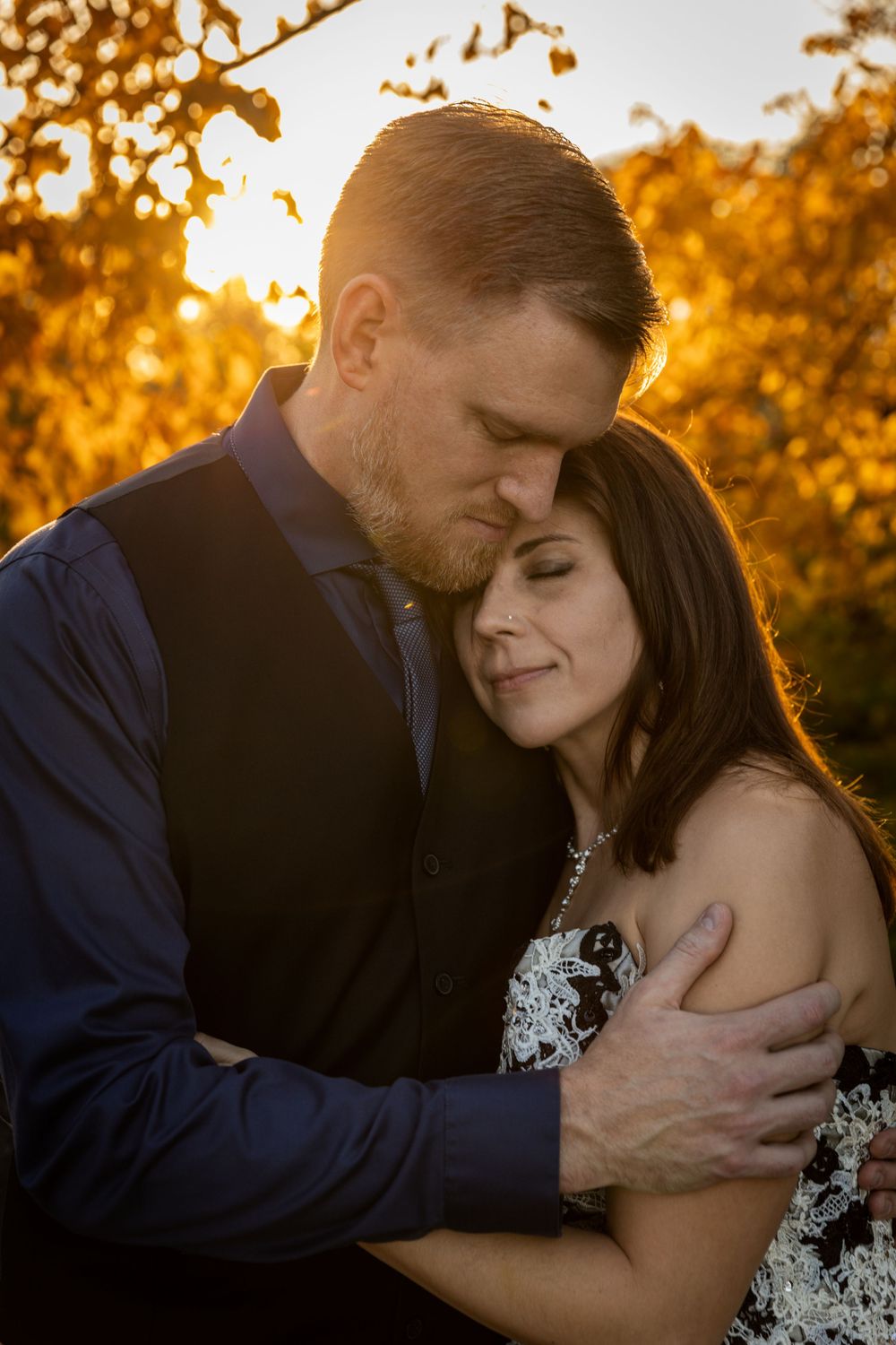 A romantic couple embraces during golden hour sunset with autumn foliage glowing warmly in the background.