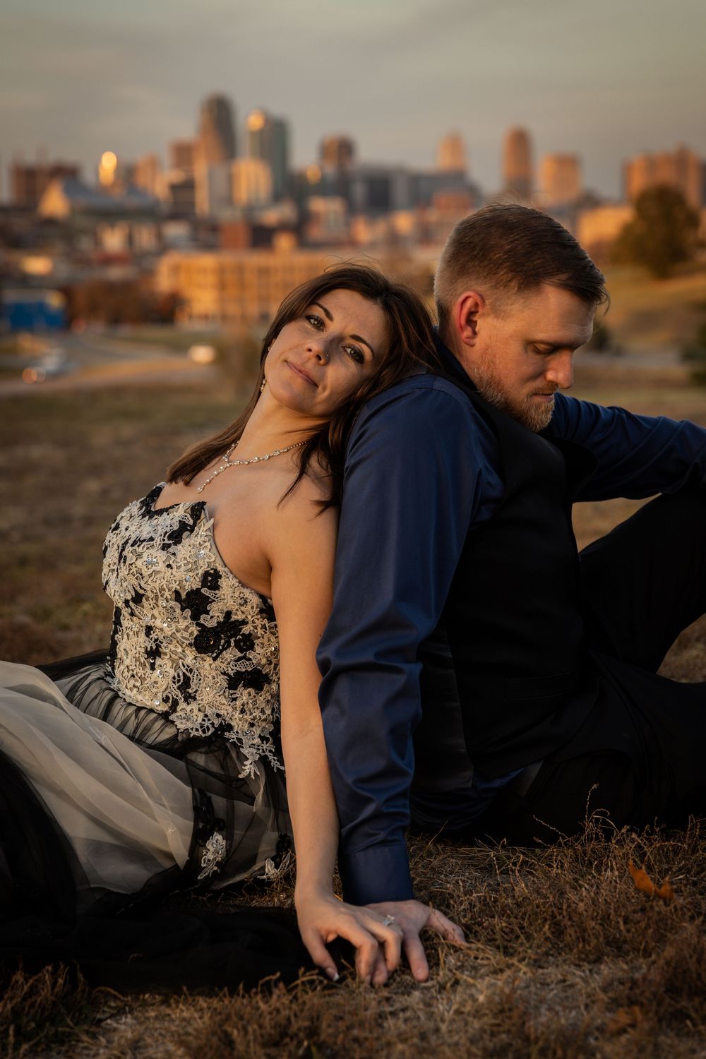A couple sits on a hill at sunset with the Kansas City skyline glowing warmly in the background.