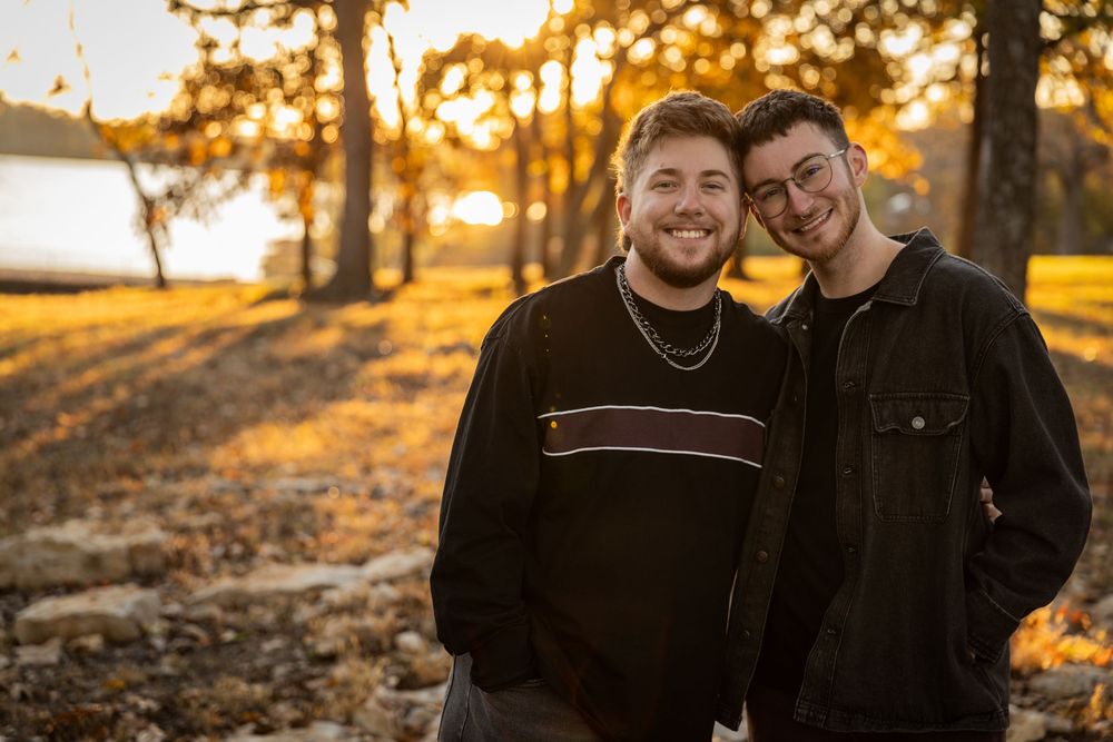 Two people stand close together smiling outdoors during golden hour sunset with trees and water in background.