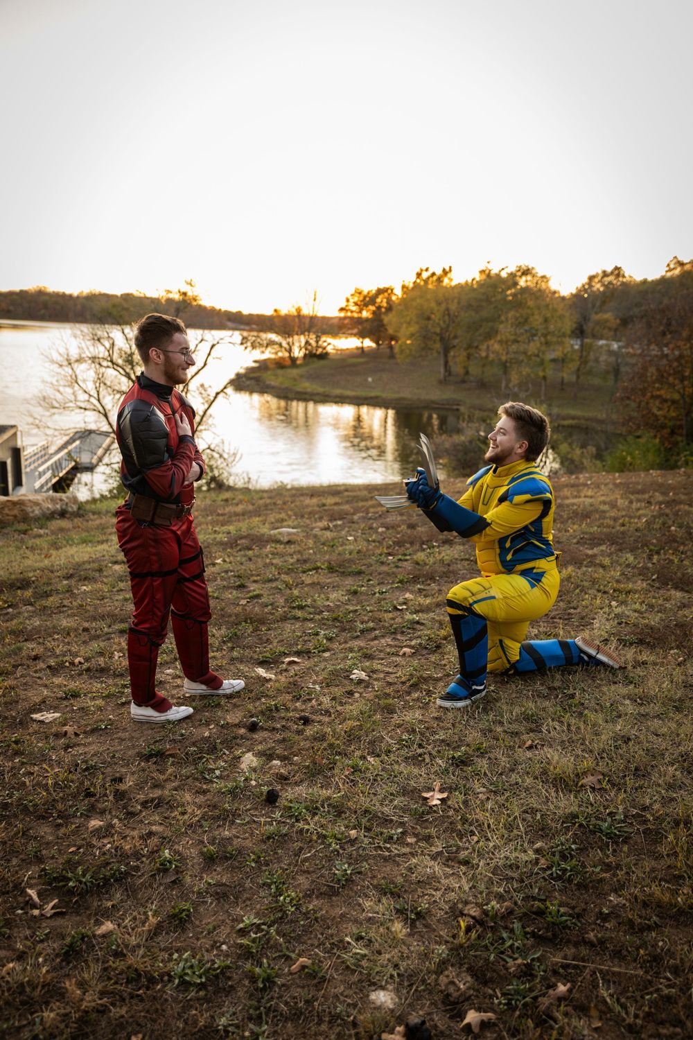 Two people in colorful winter gear playfully interact on a grassy riverbank at sunset.