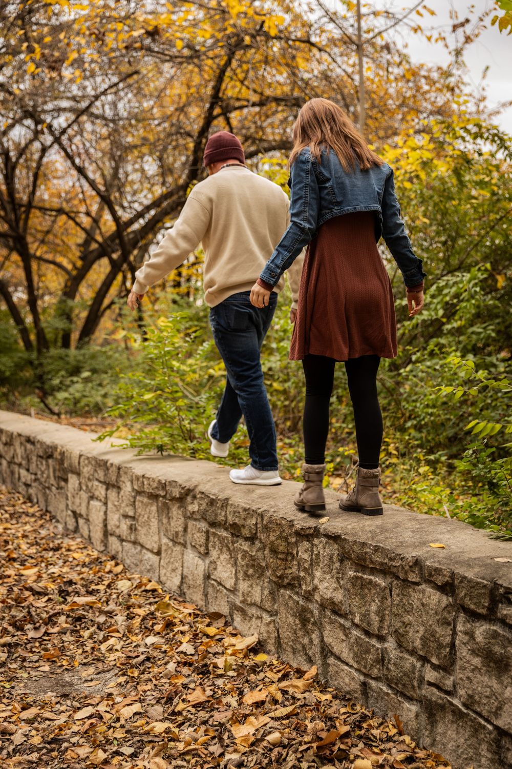 Two people walk along a stone wall in autumn with fallen leaves and golden foliage in the background.