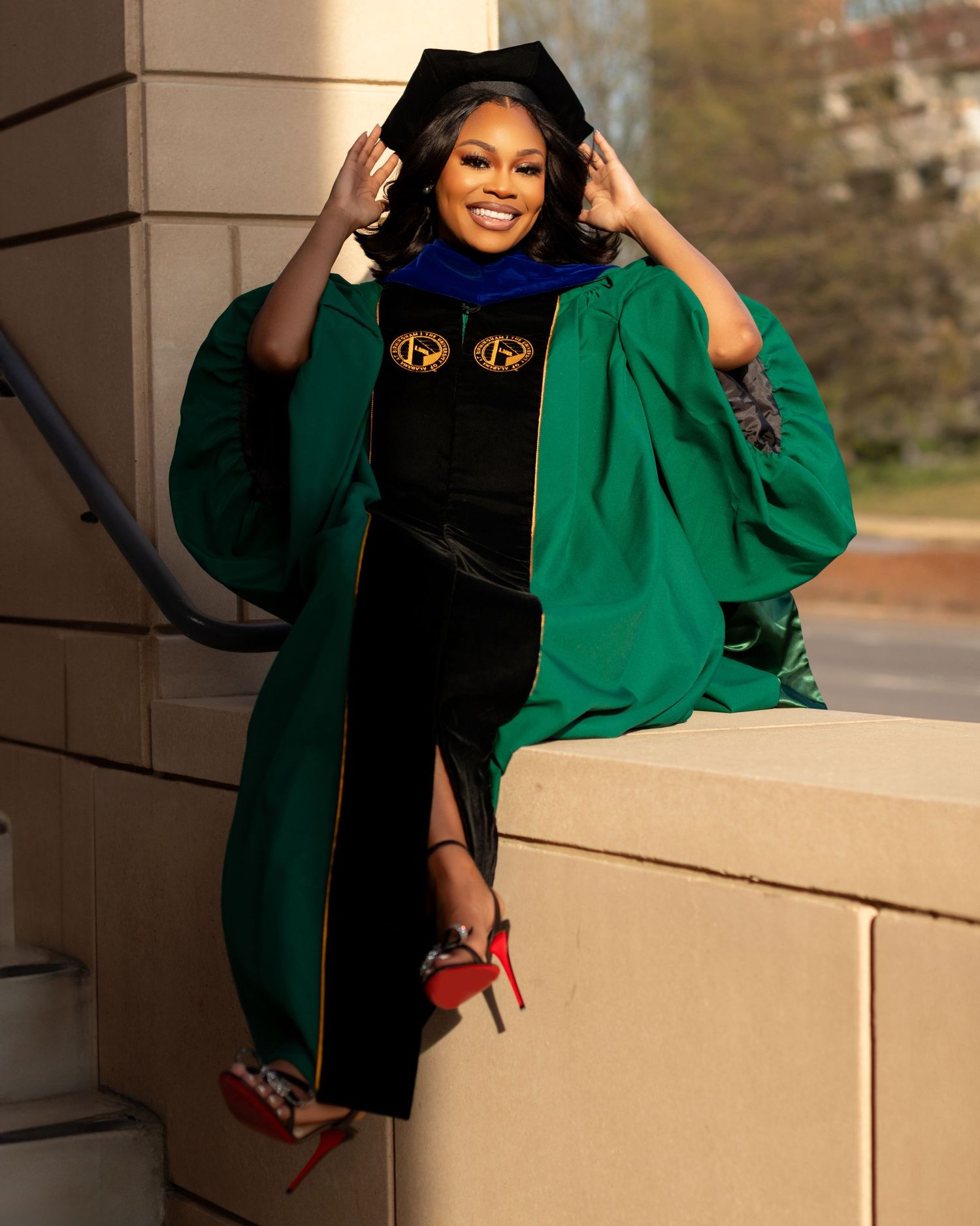 Graduate in green and black regalia poses confidently under columned archway during commencement.