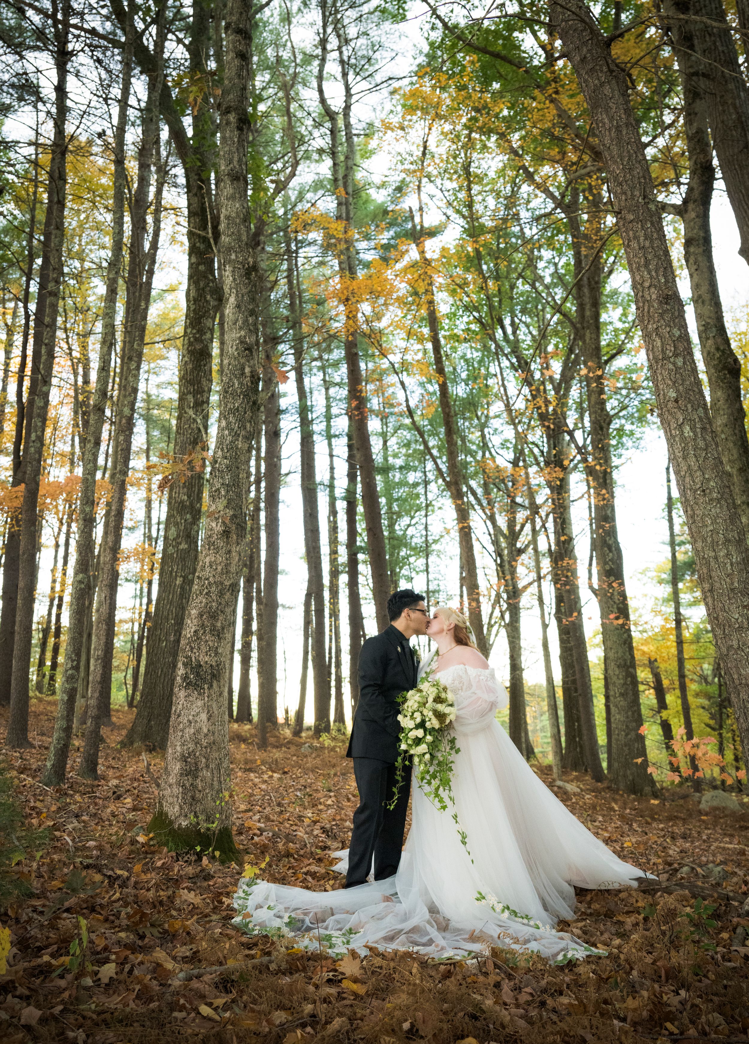 A couple in wedding attire embraces in a sunlit autumn forest with tall trees and scattered fallen leaves.