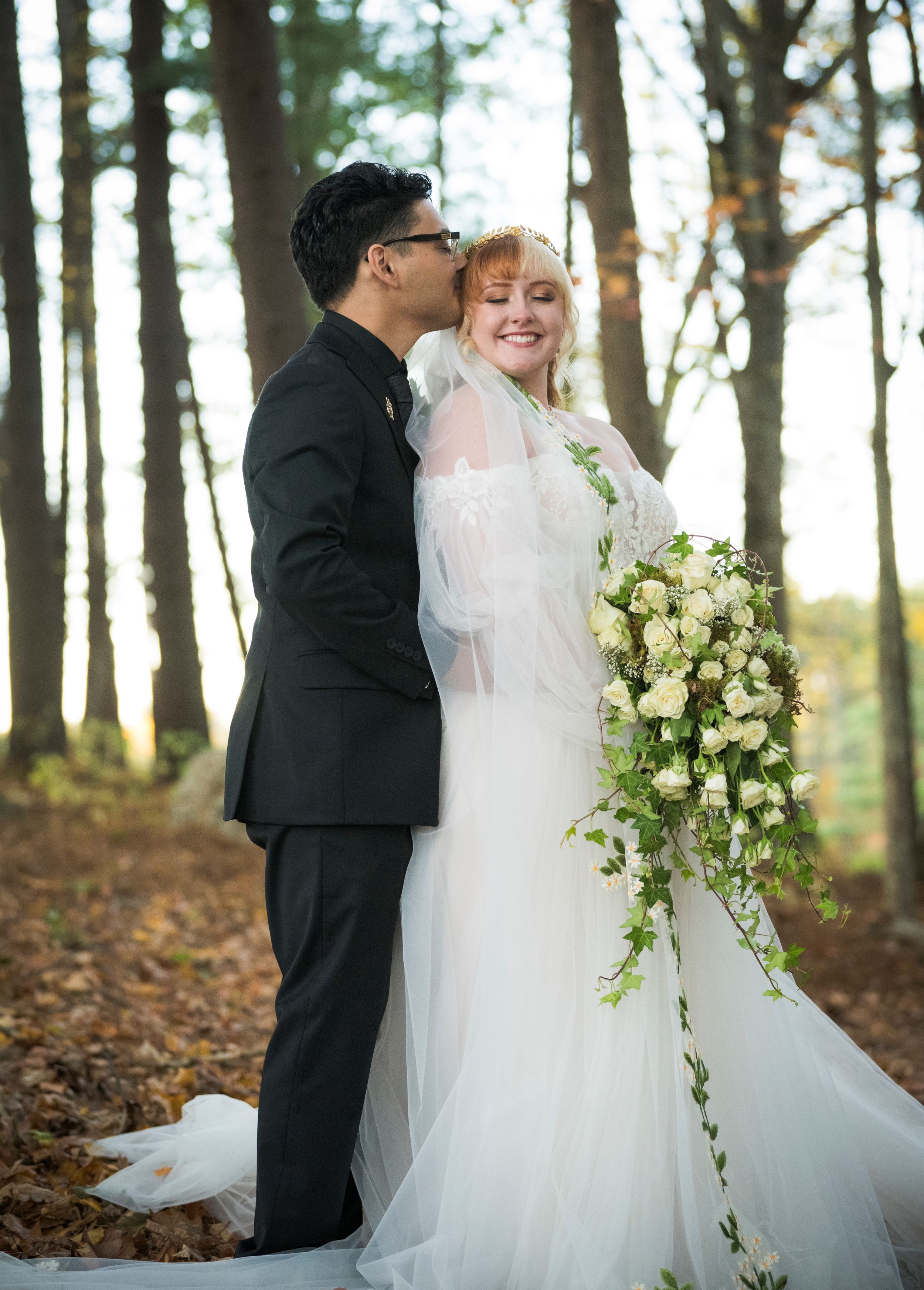 A romantic outdoor forest wedding portrait with cascading white flower bouquet and lace gown among towering pine trees.