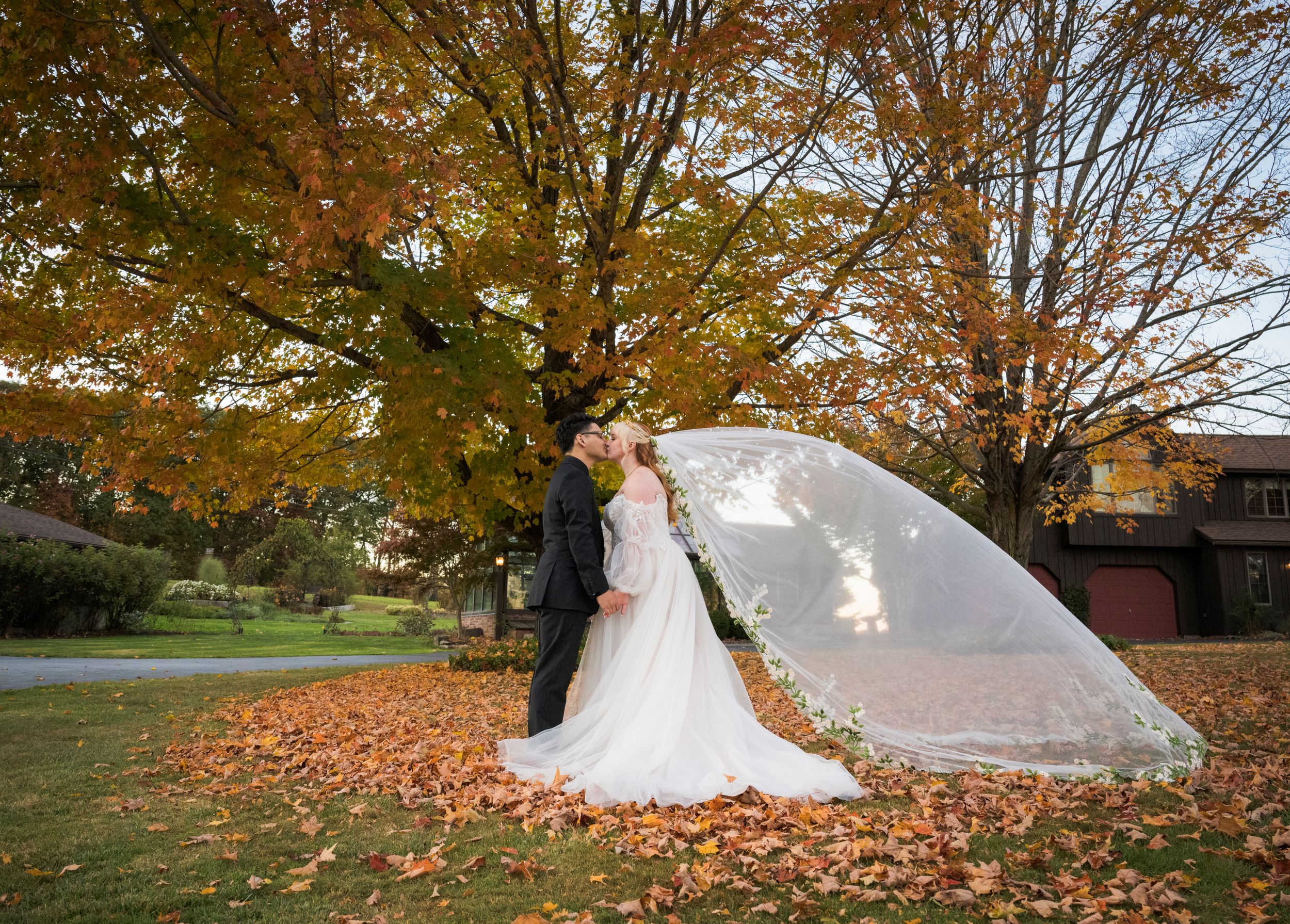 A romantic autumn wedding photo with a flowing white dress and veil against colorful fall foliage in a park setting.