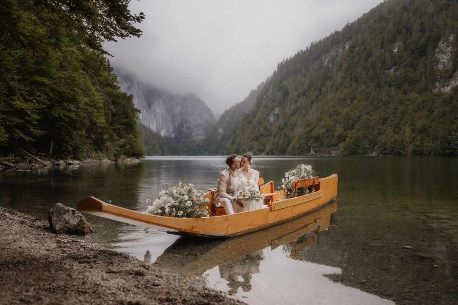 A wooden canoe rests on a misty lake surrounded by mountains and evergreen forests.
