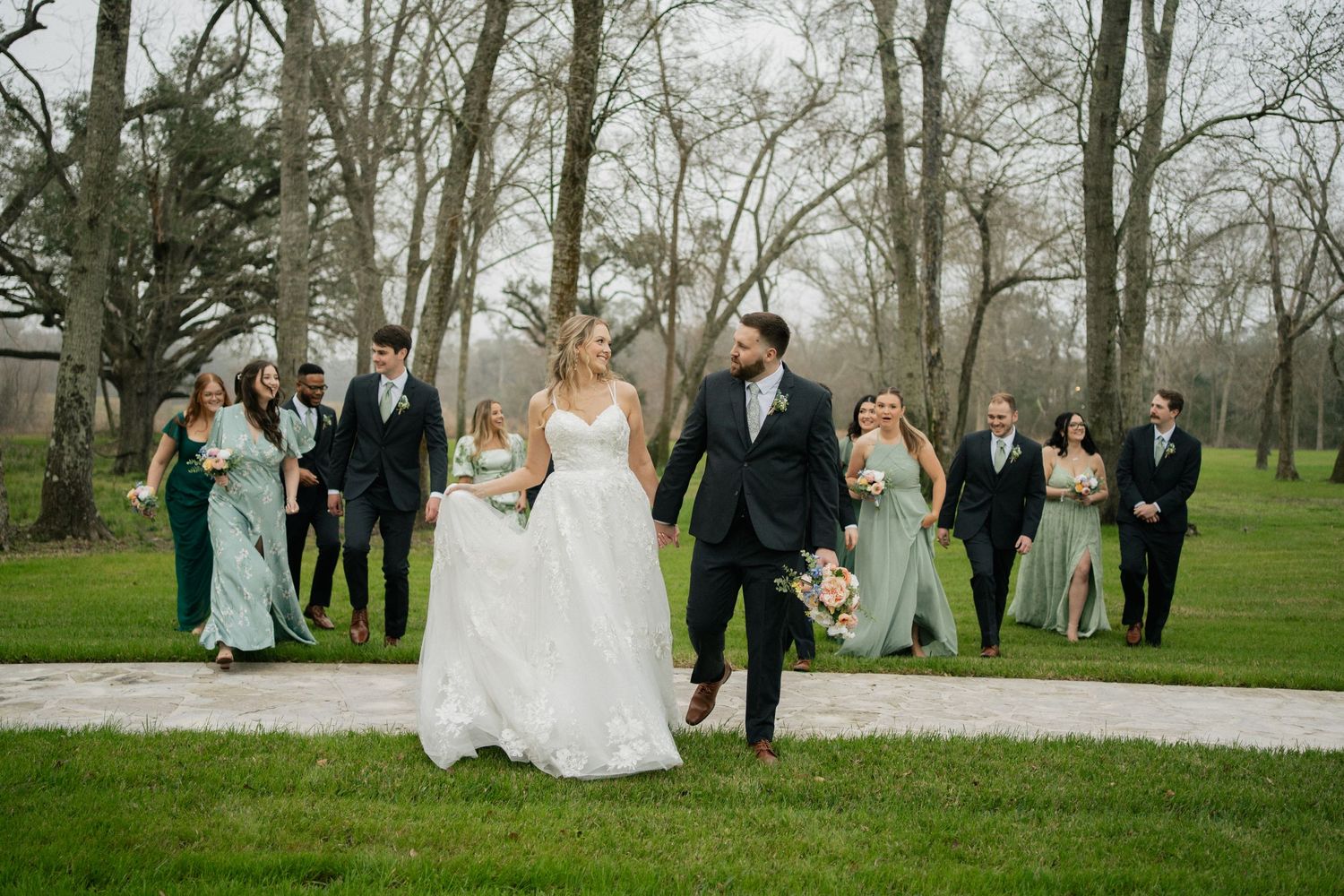 Wedding party walking on pathway through green park with bridesmaids in blue dresses and groomsmen in dark suits.