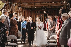 Bride walks down the aisle with her father at a rustic outdoor wedding ceremony surrounded by guests.
