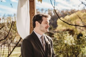 Groom in dark suit smiling outdoors at wedding ceremony with lush green trees and blue sky background.