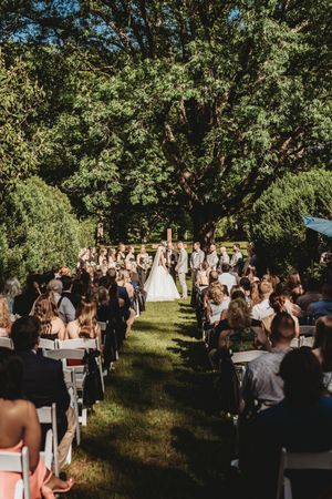 Outdoor wedding ceremony on green lawn under large trees with guests seated in white chairs along an aisle.