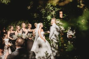 Outdoor wedding ceremony with bridesmaids holding bouquets, bride in white gown, and groom in gray suit among lush greenery.