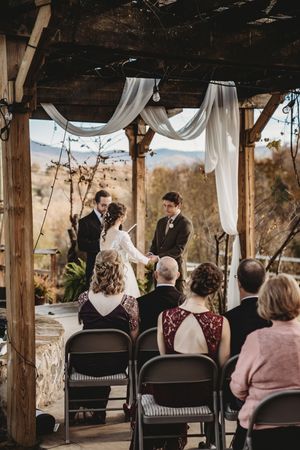 Outdoor wedding ceremony under rustic pergola with white draping, couple exchanging vows before officiant, mountain views behind.