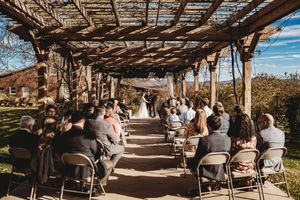 Outdoor wedding ceremony under a rustic wooden pergola with guests seated on folding chairs along a garden pathway.