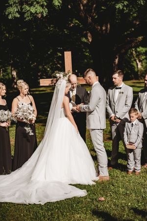 Outdoor wedding ceremony with bride in white gown and groom in gray suit exchanging vows before a wooden cross.