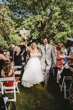 Joyful bride and groom walk down outdoor aisle after wedding ceremony, guests cheering, surrounded by lush green trees.