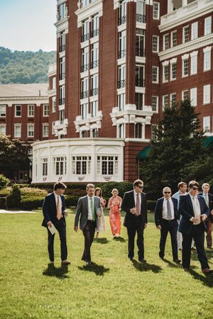 Groomsmen in dark suits walk across green lawn outside elegant red brick hotel building on sunny day.