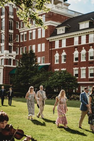 Elegantly dressed guests walk on green lawn outside a grand red brick building during an outdoor wedding reception.