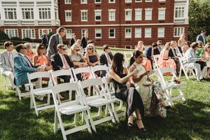 Guests seated in white folding chairs on a lawn for an outdoor wedding ceremony beside a red brick building.
