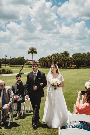 Bride in white gown walks down outdoor aisle with escort at sunny golf course wedding ceremony.