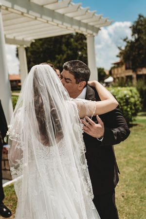 Groom in black suit kissing bride in white lace wedding gown and veil during outdoor ceremony.