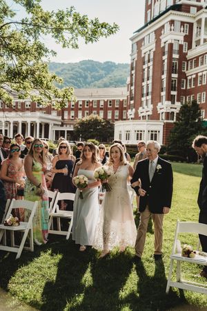 Two brides walk down outdoor aisle at elegant brick venue wedding ceremony on sunny day.