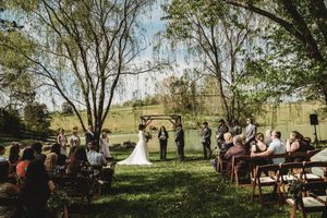 Outdoor wedding ceremony under lush green trees with guests seated on wooden chairs facing the couple at a rustic wooden arch.