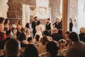 Wedding guests watch ceremony in rustic brick-walled venue with elegant white draping.